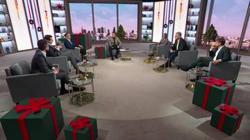 Sześciu mężczyzn siedzi w okręgu na kanapach w studiu telewizyjnym. W tle znajdują się regały z książkami i oknami z widokiem na miasto. Przed kanapami stoją zielone prezenty z czerwonymi wstążkami.