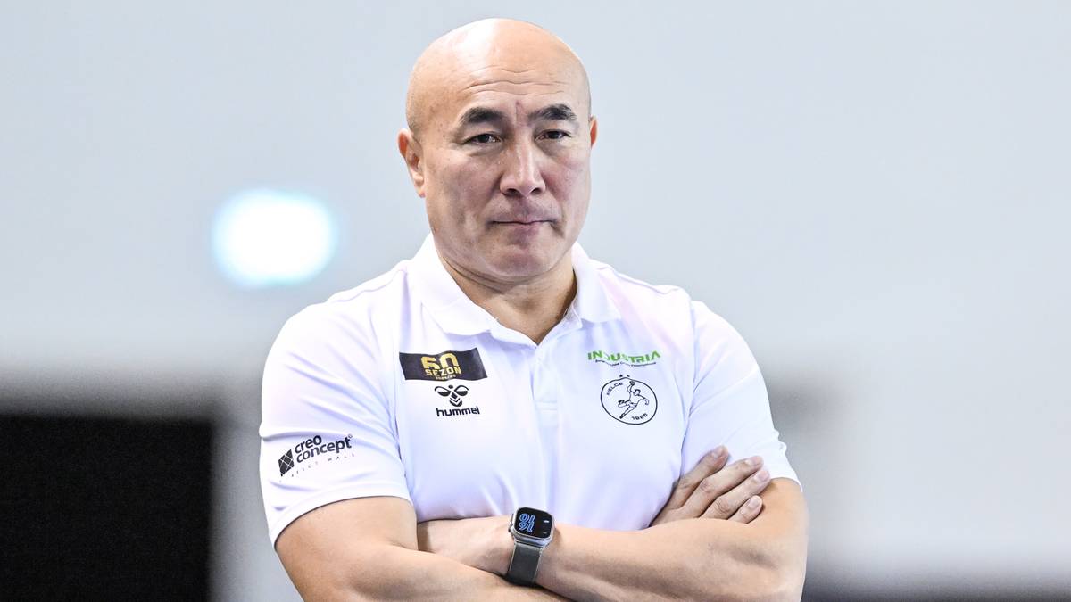 Trener drużyny sportowej w białej koszulce polo i niebieskich dżinsach stoi ze skrzyżowanymi ramionami.
