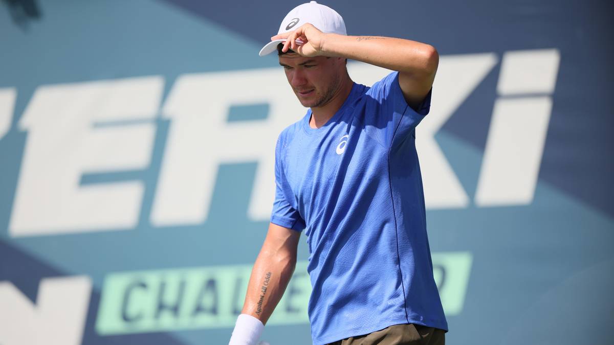 Tenisista w niebieskiej koszulce z białym logo na piersi, w białej czapce z daszkiem, poprawia czapkę dłonią przy czole. Tło stanowi niebieski ekran z białymi napisami.