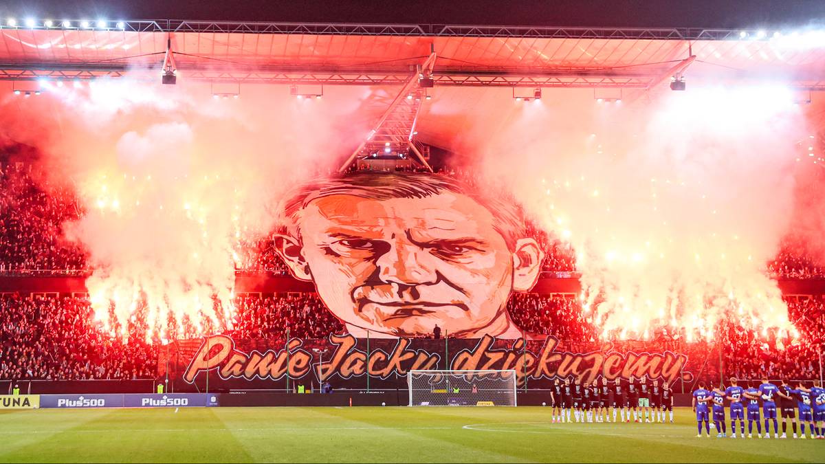 Trybuna stadionu wypełniona kibicami z flagami i racami, na środku ogromny transparent z podobizną twarzy mężczyzny i napisem &bdquo;Panie Jacku dziękujemy&rdquo;.