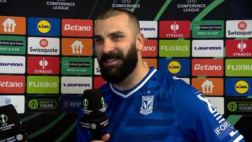 Mikael Ishak, piłkarz Lecha Poznań, w niebieskiej koszulce z herbem klubu, na tle ścianki z logotypami sponsorów.