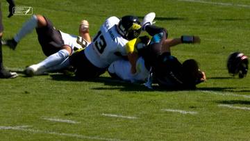 Gracze w białych i czarnych strojach rywalizują o piłkę podczas meczu futbolu amerykańskiego. Jeden z zawodników leży na ziemi, podczas gdy inny próbuje go powalić.