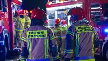 Dw&oacute;ch strażak&oacute;w w odblaskowych kurtkach z napisem "Państwowa Straż Pożarna" i kaskach, stojący na tle woz&oacute;w strażackich.