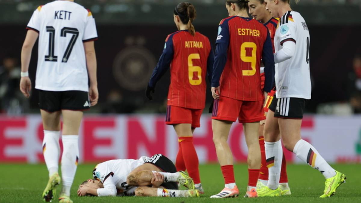 Piłkarki w białych i czerwonych strojach sportowych stoją wokół leżącej na murawie zawodniczki.