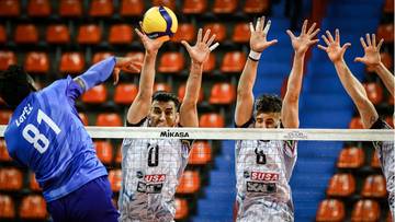 Siatkarz w niebieskiej koszulce z numerem 81 atakuje piłkę nad siatką, a trzech siatkarzy w białych strojach z numerami 0, 6 i 10 próbuje ją zablokować.