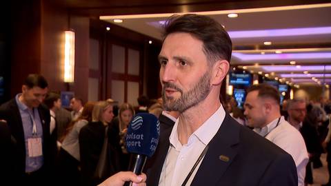 Mężczyzna z brodą w eleganckim garniturze odpowiada na pytanie z mikrofonem w ręku. W tle widać tłum ludzi na tle oświetlonych ścian i lamp. Obraz przedstawia wywiad podczas branżowego wydarzenia.