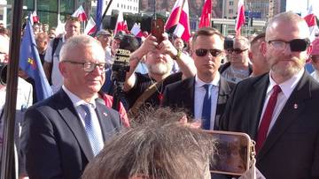 Mężczyzna w garniturze robi zdjęcie telefonem kom&oacute;rkowym na tle tłumu z flagami.