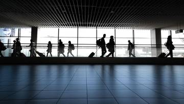 Sylwetki podróżnych z walizkami idące przez jasny, przeszklony korytarz na lotnisku.