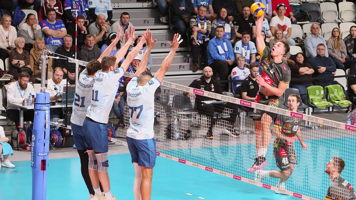 Siatkarze w akcji podczas meczu siatkówki. Jeden z graczy wykonuje wysoki skok do ataku nad siatką, próbując zdobyć punkt. Inni zawodnicy blokują lub bronią.