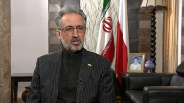 Mężczyzna w okularach i garniturze z wąsami i brodą, siedzi i m&oacute;wi, za nim flaga Iranu.