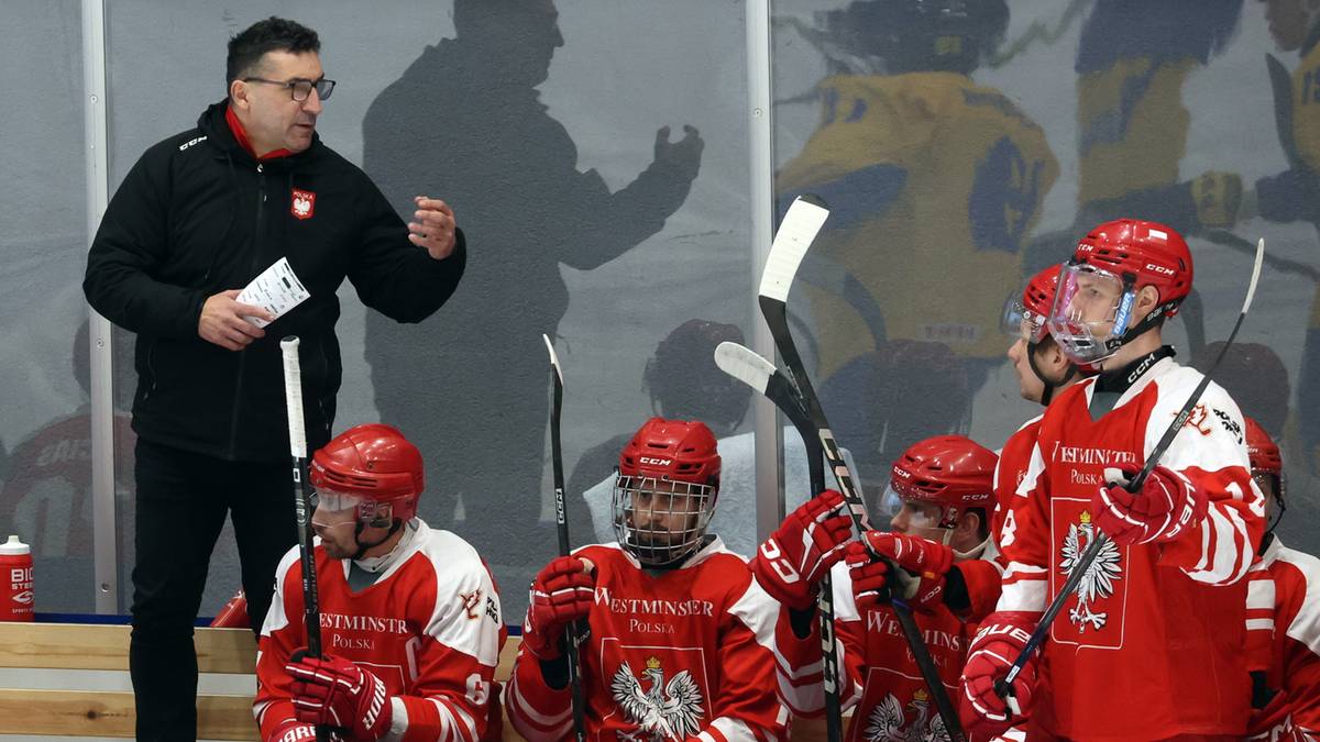 Trener w okularach przemawia do grupy zawodników hokejowych w czerwonych strojach, z innymi zawodnikami na drugim planie.