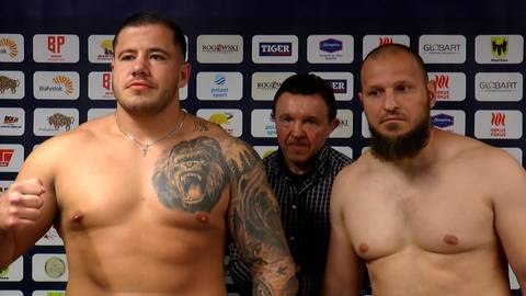 Dw&oacute;ch bokser&oacute;w stoi przed ścianą z logo sponsor&oacute;w. Na pierwszym planie po lewej stronie widoczny jest zawodnik z dużym tatuażem goryla na ramieniu, odziany w naszyjnik. Po jego prawej stronie, nieco w tle, stoi mężczyzna w kratę. Po prawej stronie, na pierwszym planie, stoi drugi bokser z brodą.