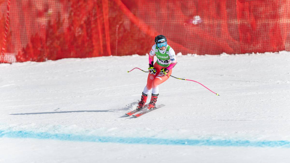 Narciarka alpejska w stroju sportowym z kaskiem i nartami zjeżdża po stoku narciarskim, z czerwonymi siatkami zabezpieczającymi w tle.