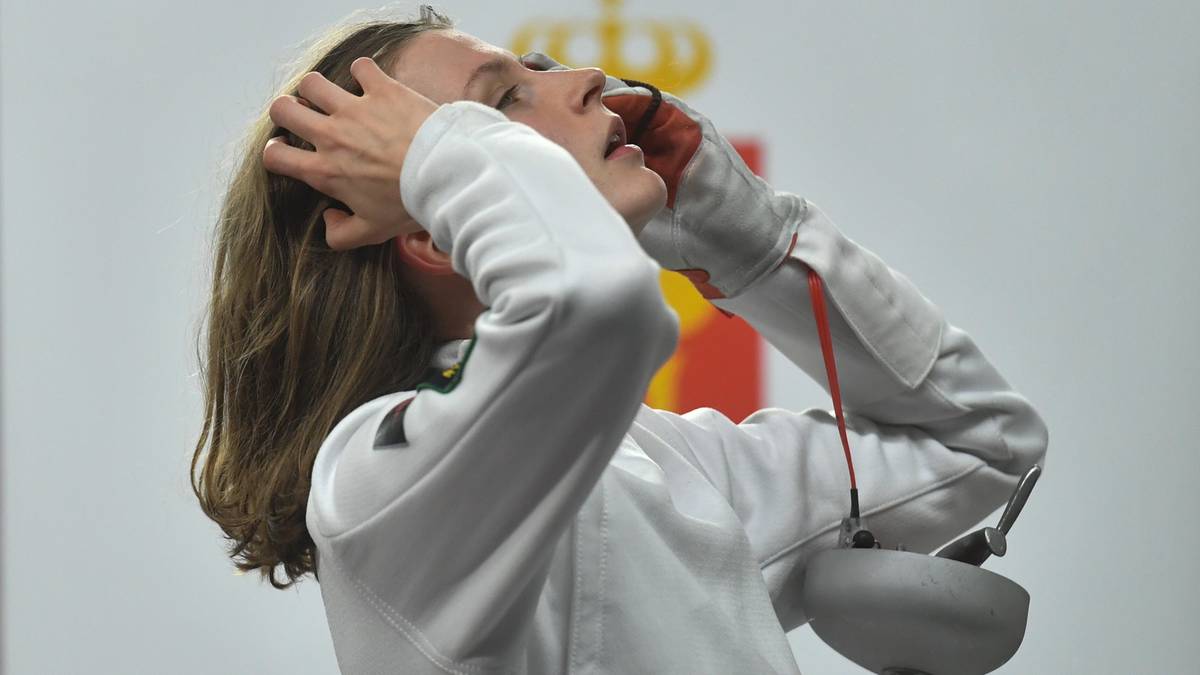 Szpadzistka w białym stroju sportowym i masce szermierczej trzymająca ręce przy głowie, wyrażająca triumf lub ulgę.