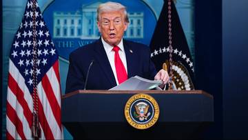 Donald Trump przemawia za m&oacute;wnicą z amerykańską flagą po lewej stronie.