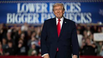 Donald Trump w garniturze i czerwonym krawacie stoi na tle banera.