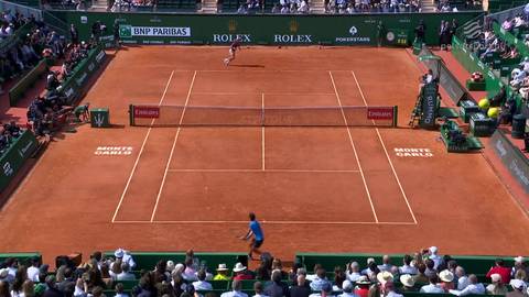 Widok z g&oacute;ry na mecz tenisa na korcie ceglanym, z dwoma zawodnikami w akcji i widzami na trybunach.