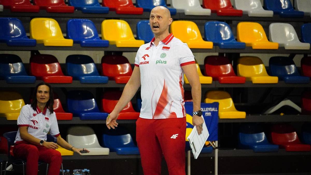 Trener siatkarzy w białej koszulce i czerwonych spodniach stoi na tle rzędu kolorowych siedzeń.