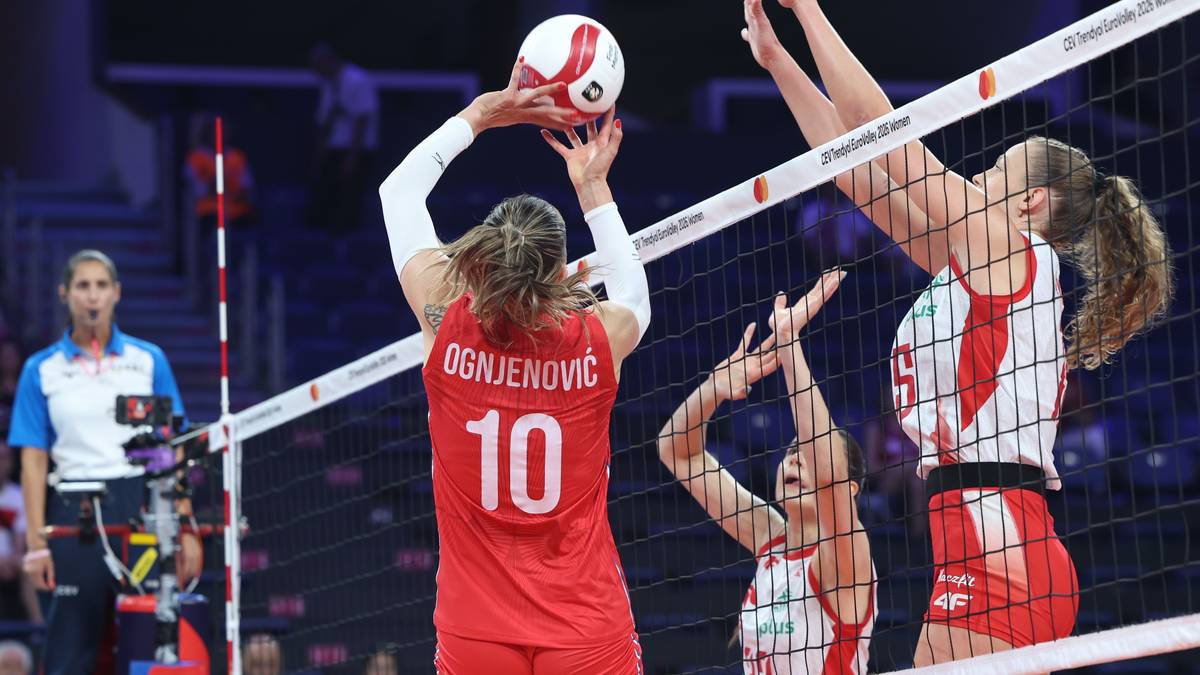 Zawodniczka w czerwonej koszulce z numerem 10 podbija piłkę do siatk&oacute;wki, podczas meczu. Dwie zawodniczki po drugiej stronie siatki pr&oacute;bują ją zablokować.