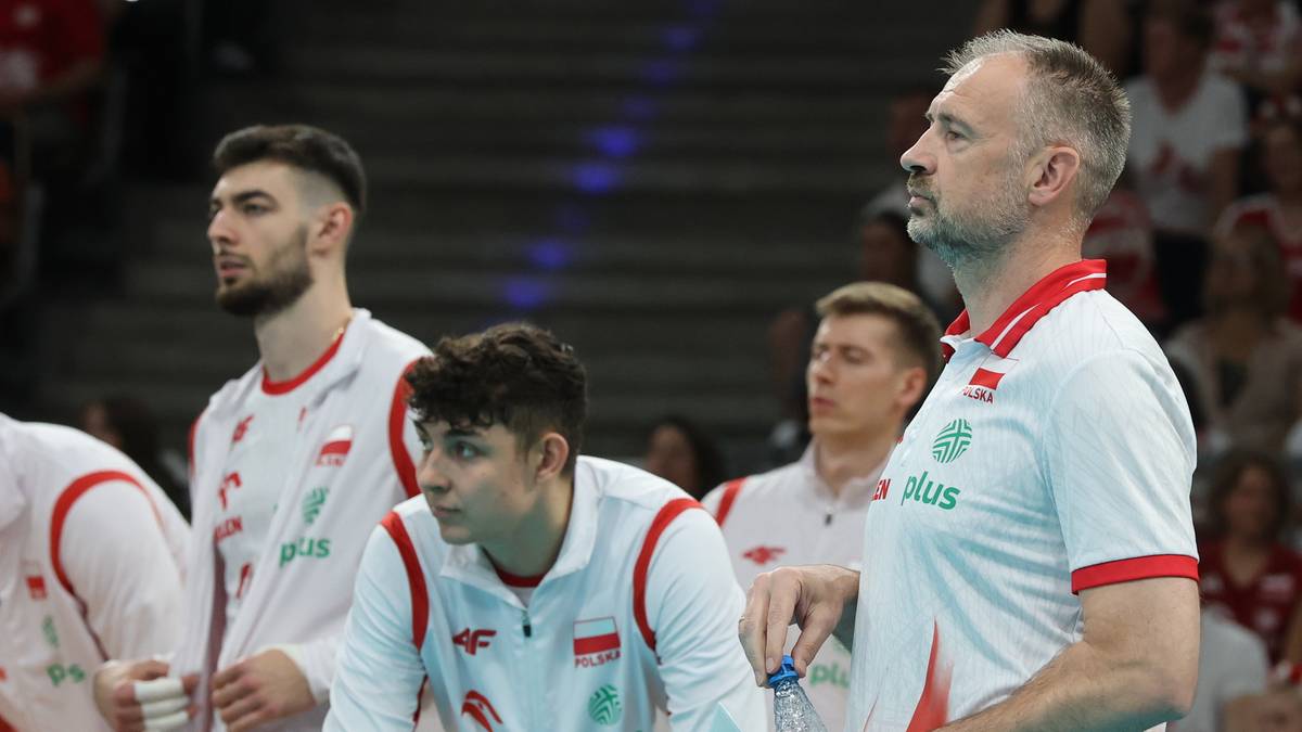 Trener siatkarzy Nikola Grbić stoi na boisku w otoczeniu młodych zawodników w białych strojach z czerwonymi elementami, z dumą patrząc w kierunku gry.