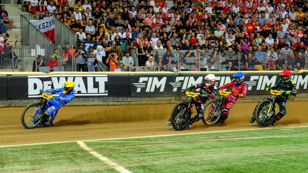 Czterech zawodników na motocyklach żużlowych ściga się po torze.