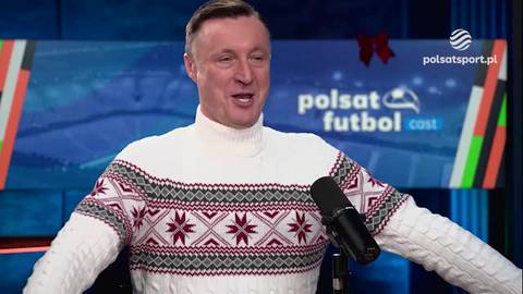 Mężczyzna w białym swetrze z czerwonym i czarnym wzorem, nosi golf. Ma mikrofon przed ustami, a w tle widoczny jest ekran z logo "Polsat Futbol Cast" i elementami dekoracyjnymi nawiązującymi do świąt.