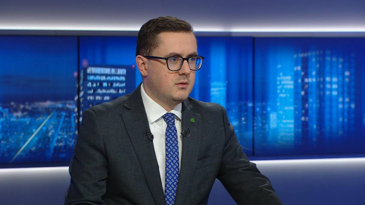 Mężczyzna w garniturze i okularach siedzi przed studiem telewizyjnym.