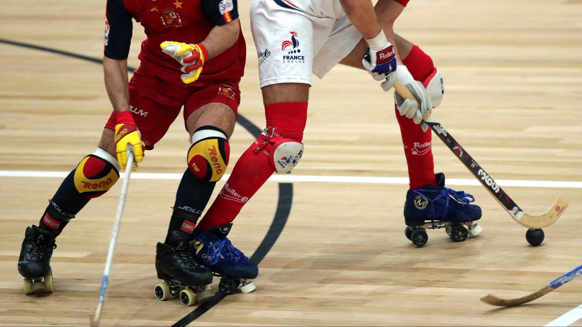 Dwóch zawodników grających w hokeja na rolkach, jeden w czerwonym stroju, drugi w białym stroju z francuskimi symbolami. Obaj mają kije hokejowe i jadą na rolkach po drewnianej nawierzchni.