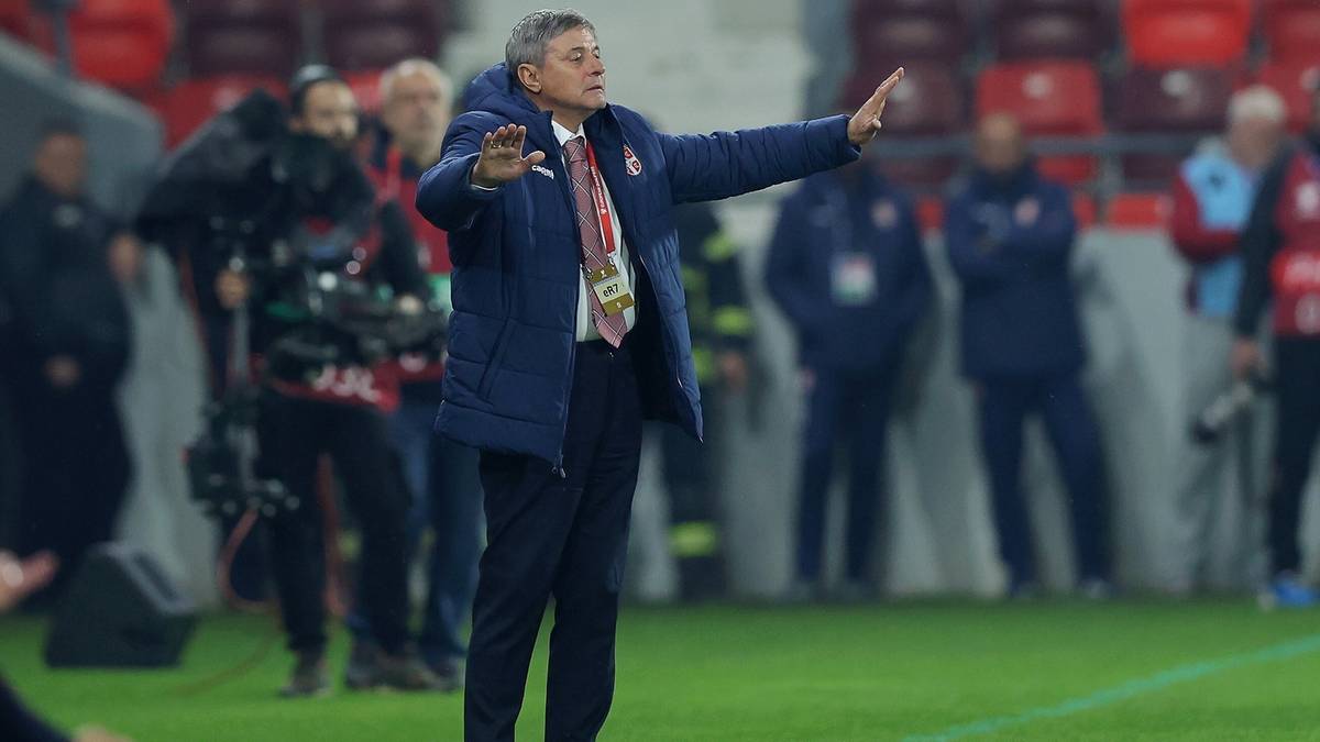 Mężczyzna w niebieskiej kurtce, spoglądający w bok z podniesioną ręką, gestykulujący na tle stadionu.