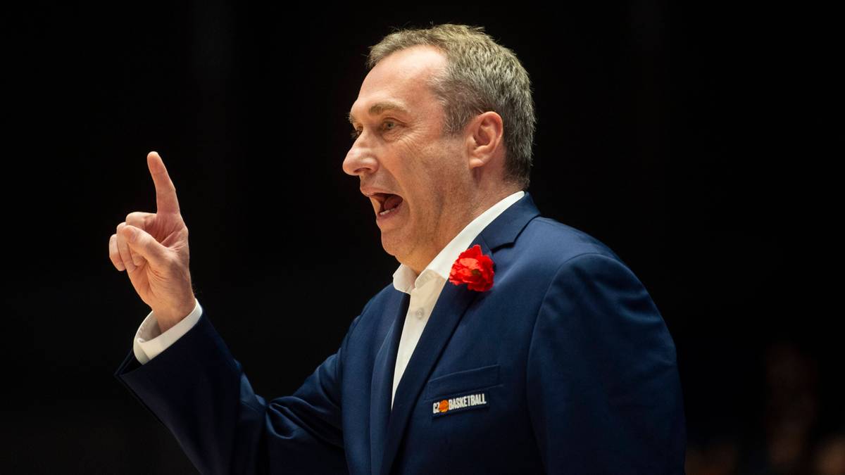 Mężczyzna w garniturze, prawdopodobnie trener koszykówki, podnosi palec wskazujący, wskazując lub przemawiając.