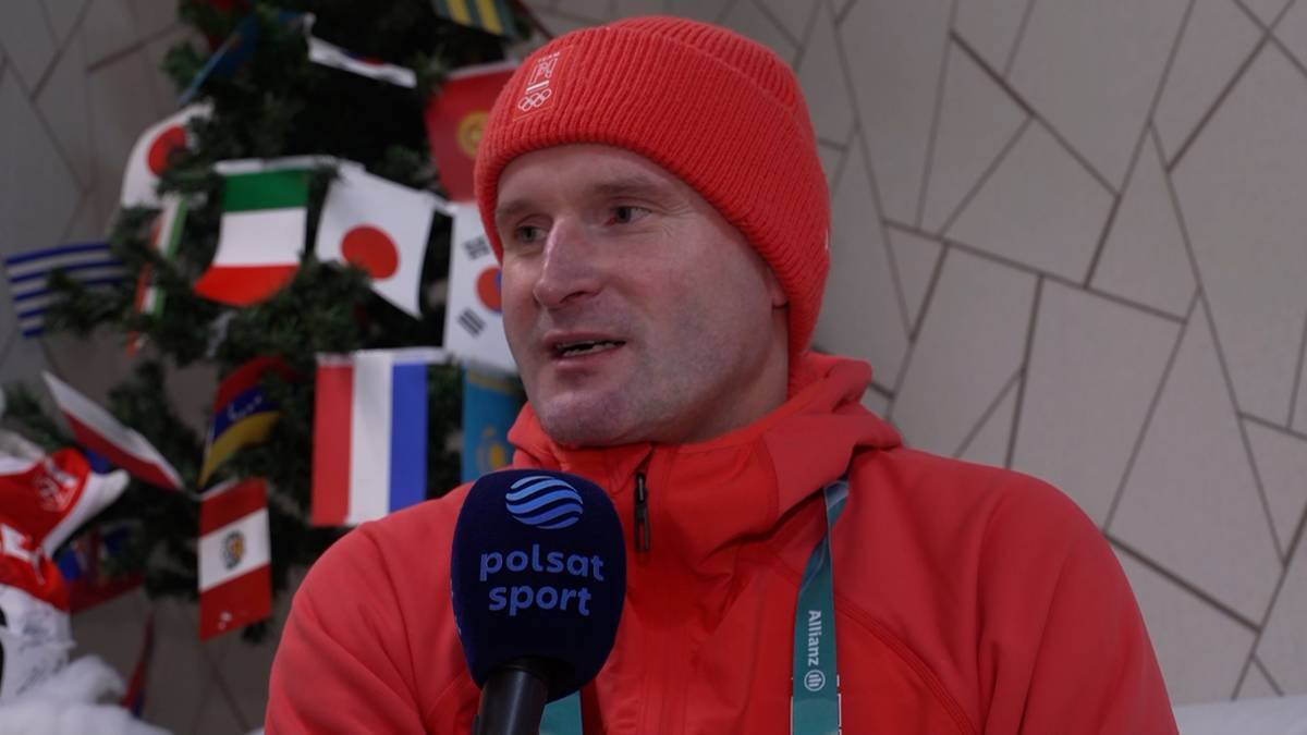 Mężczyzna w czerwonej czapce i bluzie trzyma mikrofon Polsat Sport. W tle widoczna choinka z flagami.