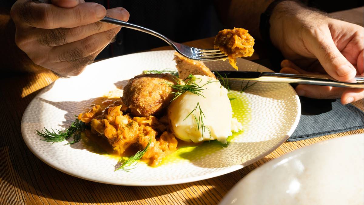 Na białym talerzu znajduje się porcja potrawy, na kt&oacute;rą składają się klopsy w gęstym sosie, puree ziemniaczane oraz zielona dekoracja. Osoba nabiera widelcem kawałek klopsa z sosem.