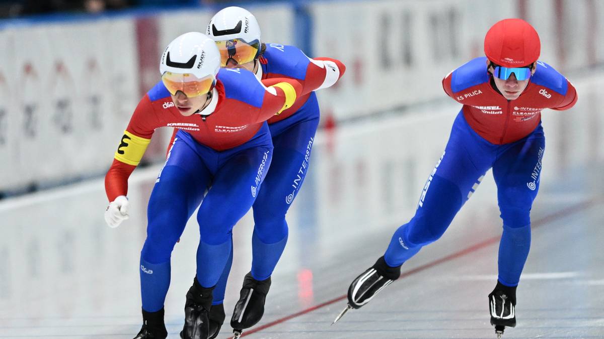 Trzech łyżwiarzy szybkich w czerwono-niebieskich strojach sportowych, rywalizujących na lodzie.