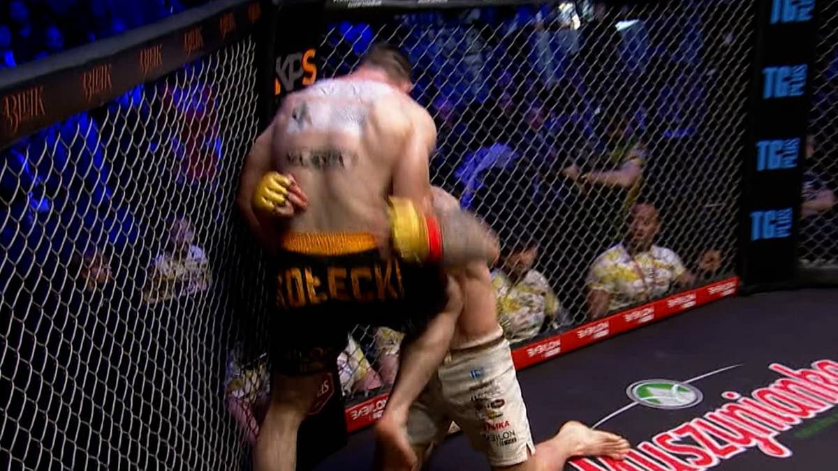 Dwóch zawodników MMA walczy w klatce, jeden z nich klęczy na macie.