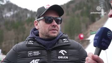 Michal Doleżal, trener polskich skoczk&oacute;w narciarskich, w czapce z daszkiem i okularach przeciwsłonecznych, udziela wywiadu. Na jego kurtce widoczne są loga sponsor&oacute;w.