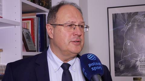 Marian Kmita w okularach, ubrany w garnitur, udziela wywiadu przed mikrofonem.