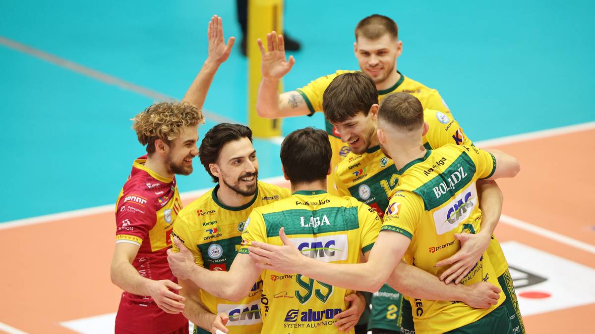 Grupa siatkarzy w ż&oacute;łtych koszulkach z numerami na plecach, przytulająca się na boisku siatkarskim. Wszyscy są uśmiechnięci i wydają się świętować zwycięstwo lub udaną akcję.