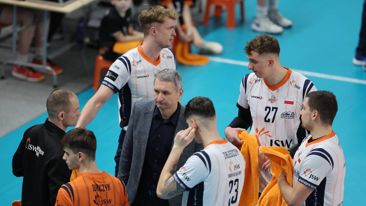 Siatkarze Jastrzębskiego Węgla w strojach sportowych dyskutują z trenerem na boisku.