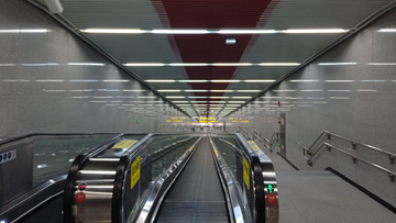 Długi korytarz stacji metra z ruchomymi schodami i schodami po prawej stronie. Sufit jest wyłożony panelami z oświetleniem i ozdobiony czerwonym wzorem.