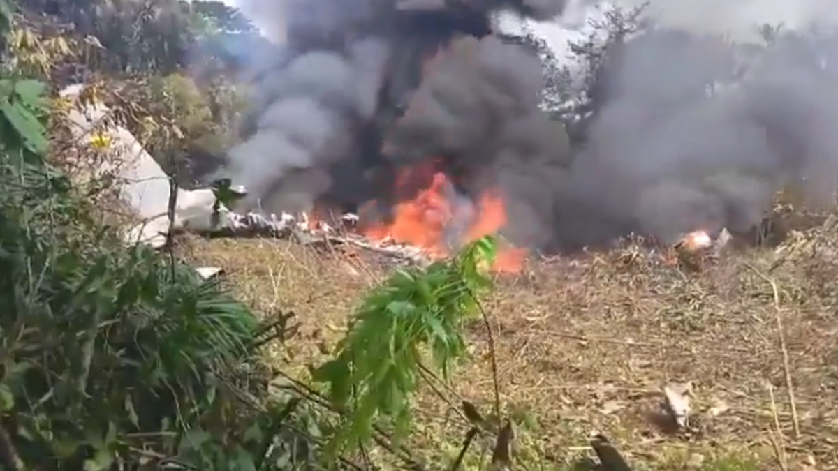 Wrak samolotu transportowego C-130 w płomieniach, z kt&oacute;rego unosi się gęsty dym na tle zielonej roślinności.