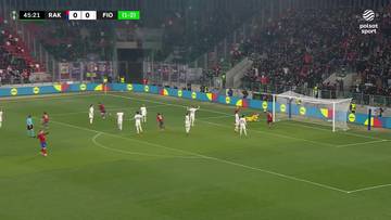 Na boisku piłkarskim trwa mecz pomiędzy drużynami Rak&oacute;w Częstochowa i Fiorentina. Na tablicy wynik&oacute;w widnieje wynik 1:0 dla Rakowa. Widać zawodnik&oacute;w na murawie, część z nich świętuje zdobycie bramki. Na pierwszym planie zielona murawa i bramka. W tle rozległe trybuny wypełnione widzami.