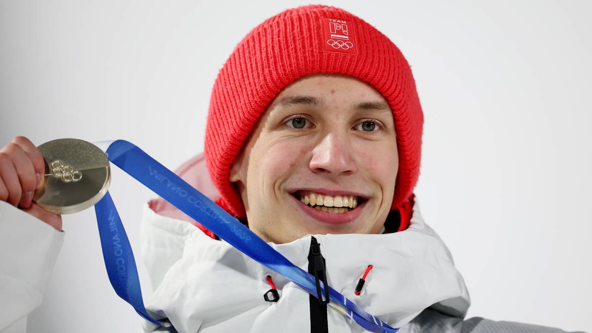 Młody mężczyzna w czerwonej czapce i kurtce narciarskiej, trzymający srebrny medal na niebieskiej wstążce.