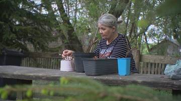 Kobieta z siwymi włosami siedzi przy drewnianym stole na zewnątrz, a przed nią znajdują się doniczki i niebieski kubek. Pali papierosa.
