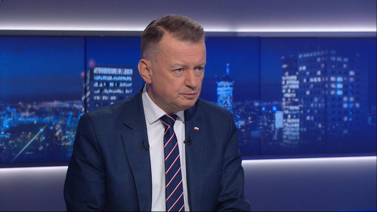 Mężczyzna w garniturze i krawacie stoi przed ekranem z nocnym widokiem miasta.