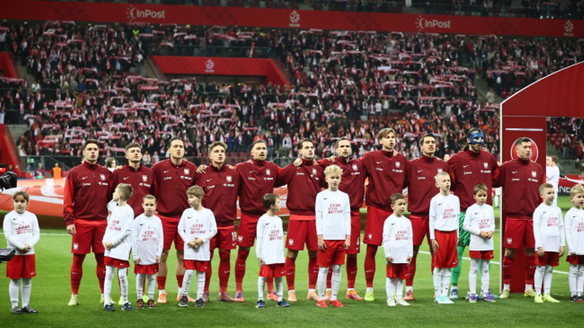 Reprezentacja Polski w strojach meczowych, z dziećmi w białych strojach, stoi na boisku piłkarskim.