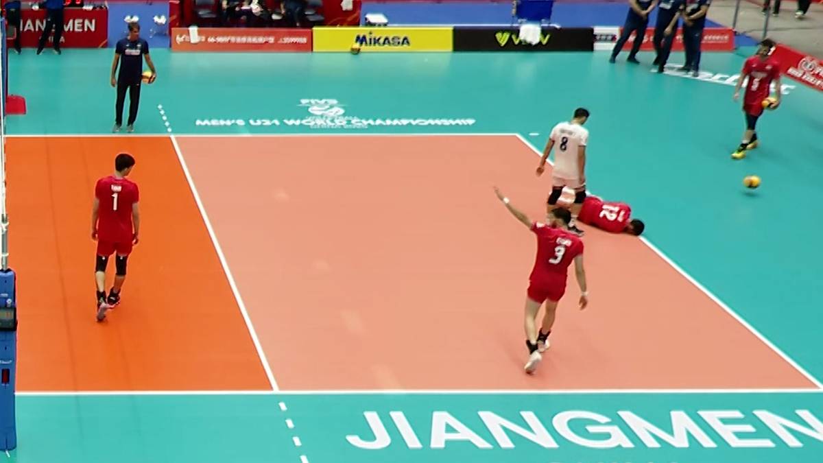 Siatkarz w czerwonej koszulce leży na boisku, prawdopodobnie kontuzjowany, podczas rozgrzewki przed meczem. Inni zawodnicy w czerwonych koszulkach i sędzia obserwują sytuację.