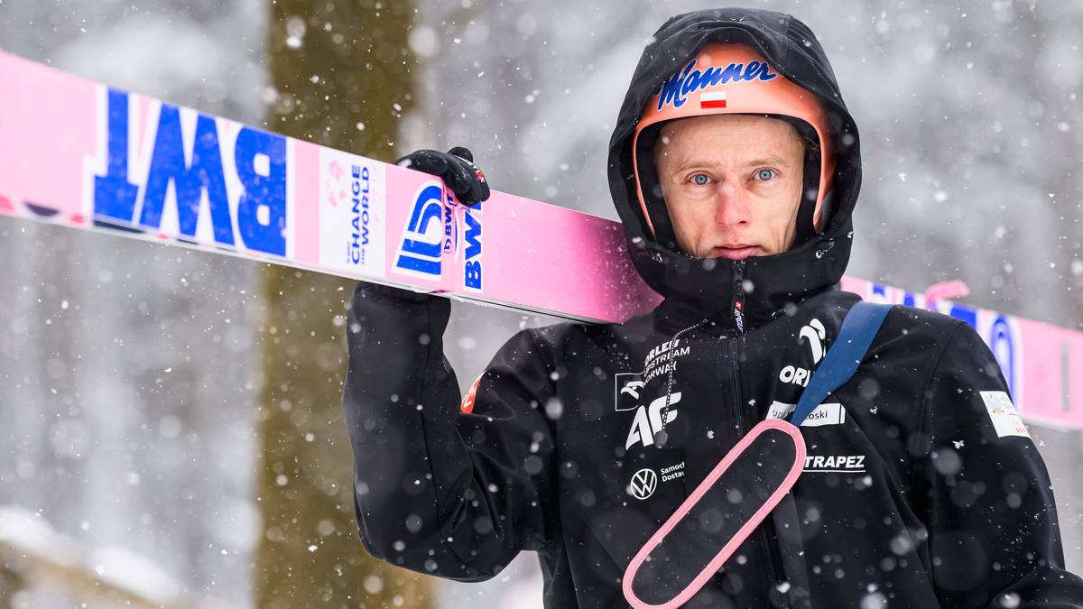 Skoczek narciarski w kasku i kurtce narciarskiej trzyma narty na ramieniu.
