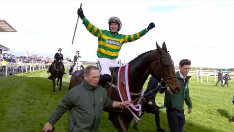 Jeździec w zielono-ż&oacute;łtym stroju triumfalnie unosi rękę w g&oacute;rę, siedząc na koniu podczas wyścigu.