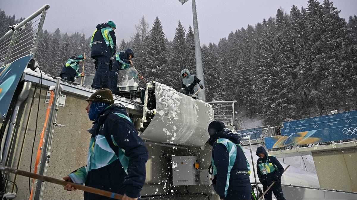 Osoby w niebieskich kurtkach i czapkach pracują przy śniegu i lodzie na tle zalesionych wzg&oacute;rz i obiektu sportowego.