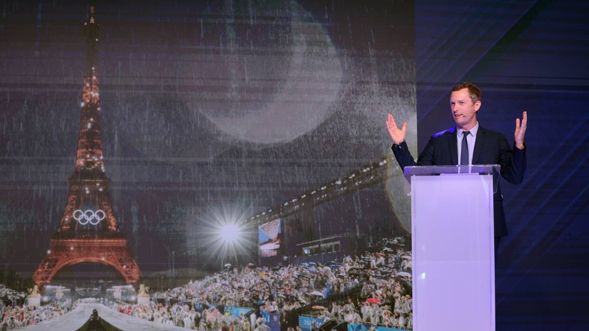 Mężczyzna przemawia za mównicą na tle ekranu z wieżą Eiffla i symbolem igrzysk olimpijskich.